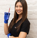 Person wearing a black scrub top and blue gloves holding a syringe against a light brick wall.