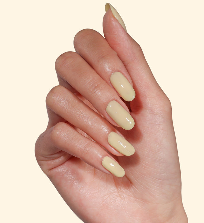 A hand with neatly manicured nails painted in LONDONTOWN’s Banana Pudding, a glossy pale yellow polish, posed against a plain light background.