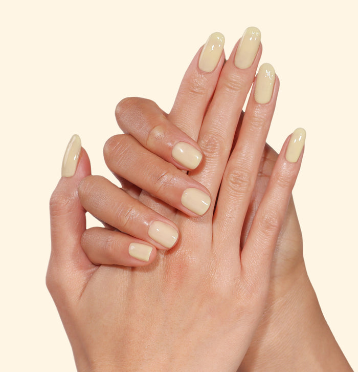 Two hands with neatly manicured nails painted in LONDONTOWN’s Banana Pudding, a pale, mellow yellow polish, set against a plain light background.