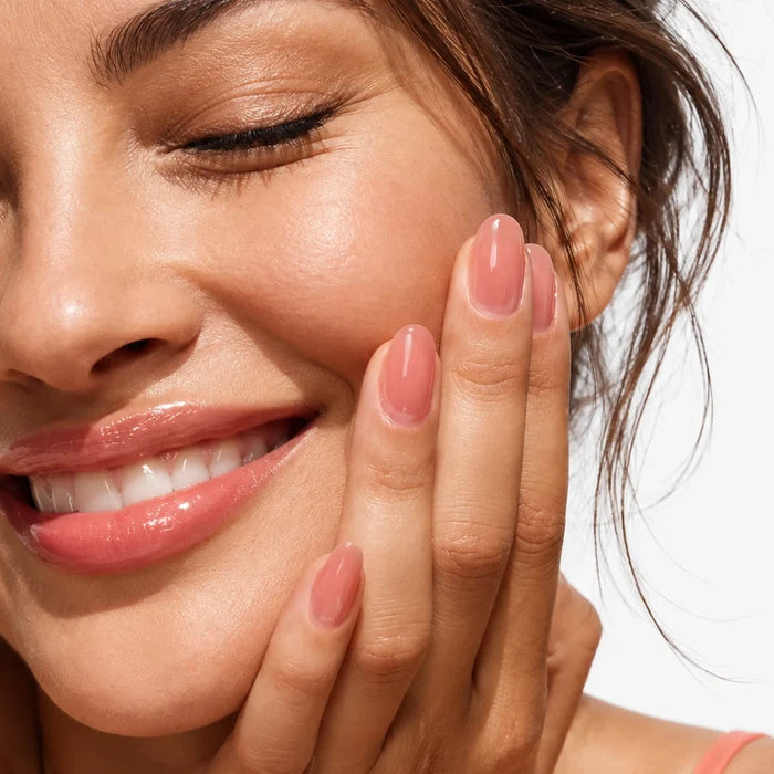 Close-up of a smiling woman with glowing skin and glossy pink fingernails painted with Bio Peptide Nail Glow resting her hand against her cheek.
