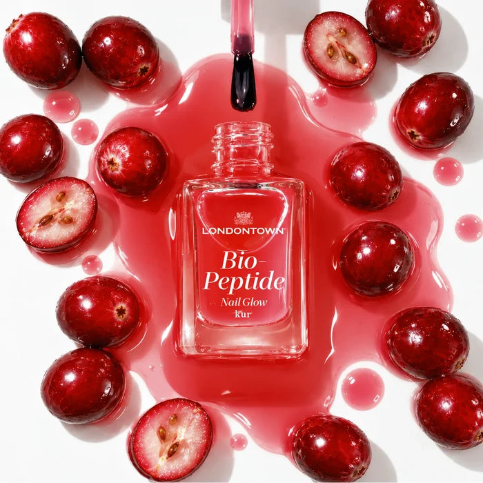 A bottle of LONDONTOWN Bio Peptide Nail Glow surrounded by fresh cranberries and a pool of translucent red liquid, evoking a dual-peptide glow for radiant skin.