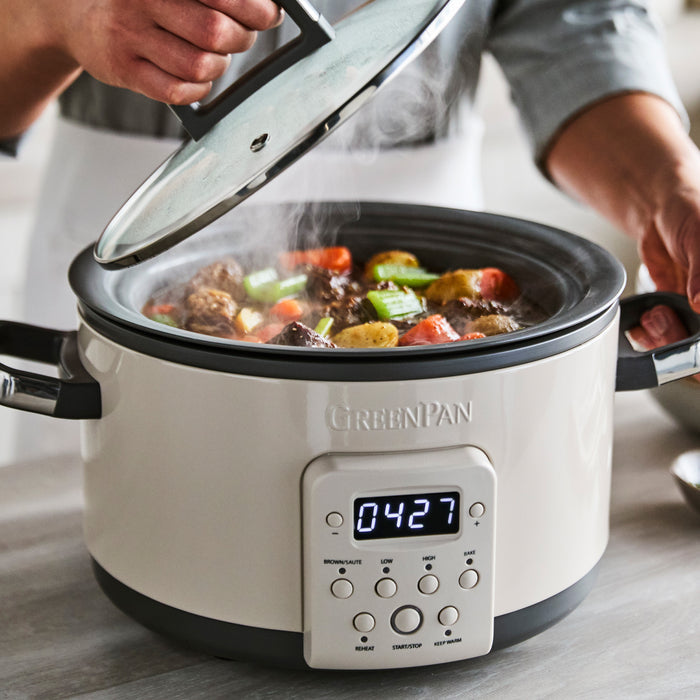 Hand lifting lid from macadamia-colored 4-quart slow cooker with steaming stew visible inside.