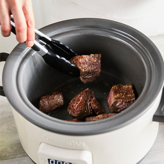 Browning beef with black tongs in a macadamia-colored 4-quart slow cooker with in-pot browning feature.