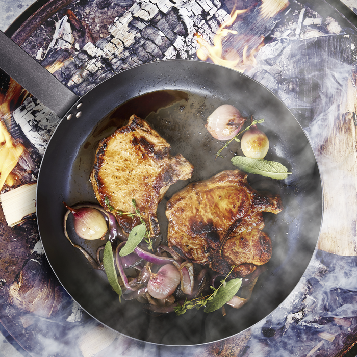 Pork chops and onions cooking in a black carbon steel pan from a 5-piece set.
