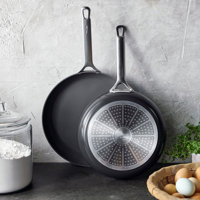 Two hard anodized frypans, 10 and 12, on a kitchen counter with flour, eggs, and fresh herbs nearby.