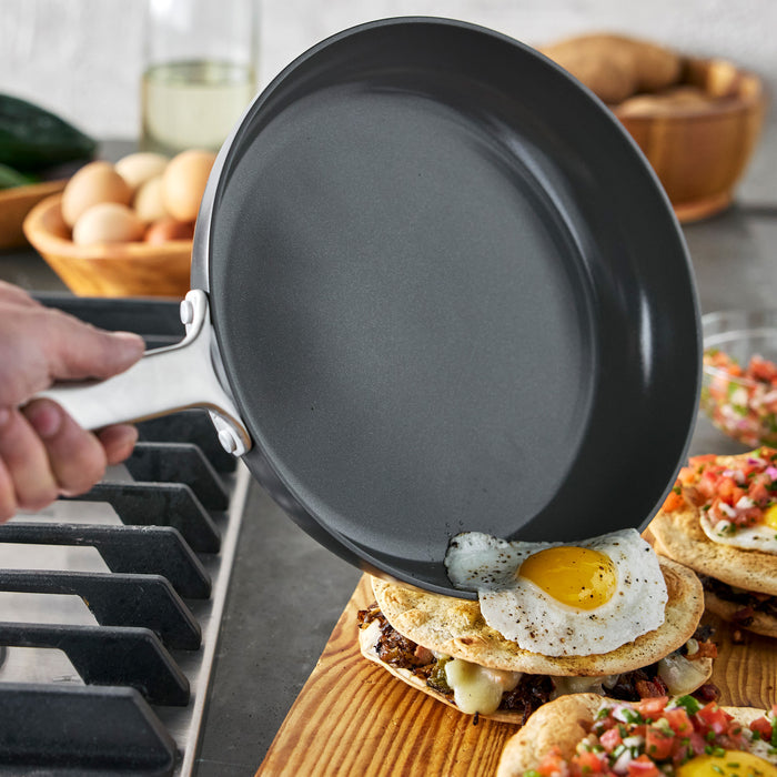 Hand sliding a fried egg from a stainless steel nonstick frypan onto food on a wooden board by the stove.