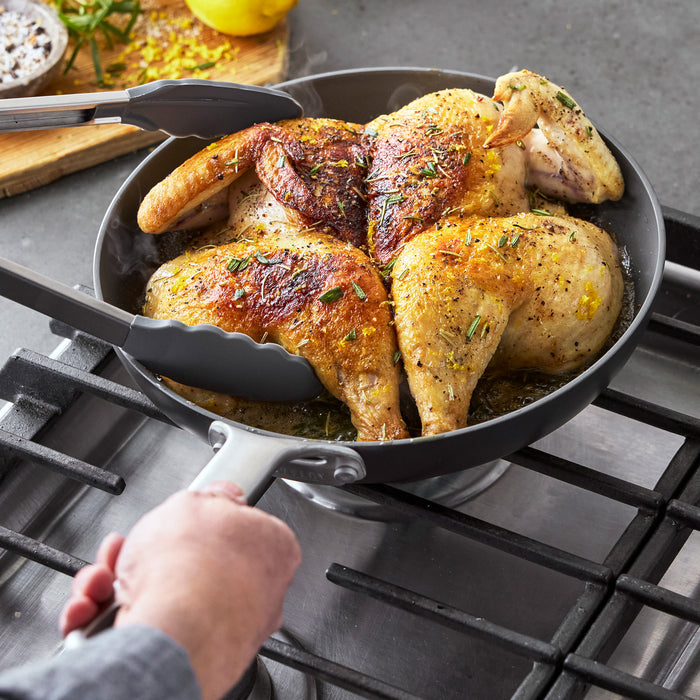 Chicken being cooked with tongs in a black hard anodized GreenPan Chef Series frypan on a stovetop.