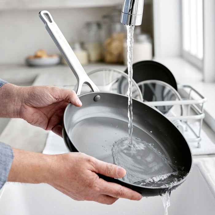 Two hard anodized frying pans being rinsed at a kitchen sink, part of a 10 and 12 frypan set.