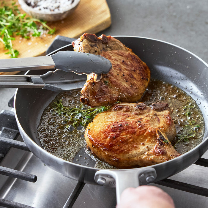Two pork chops cooking with herbs in a black Bobby Flay by GreenPan Chef Series hard anodized pan on a stovetop.