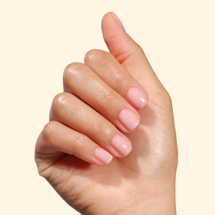 A hand with neatly manicured nails painted in LONDONTOWN’s Confetti Cake, a sweet pastel pink shade, is shown against a plain beige background.