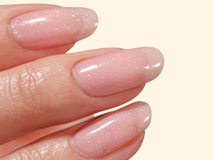 Close-up of three fingers with neatly manicured nails painted in LONDONTOWN’s Confetti Cake—a sweet, pastel pink polish featuring a subtle confetti sparkle—against a light background.