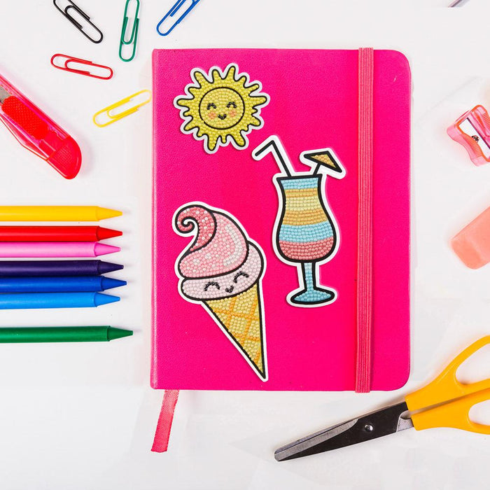 A pink notebook with Diamond Dotz Sunshine stickers and colorful designs sits among Diamond Dotz, colored pencils, paper clips, scissors, a sharpener, and an eraser—ideal for any kids’ craft kit on a bright white surface.