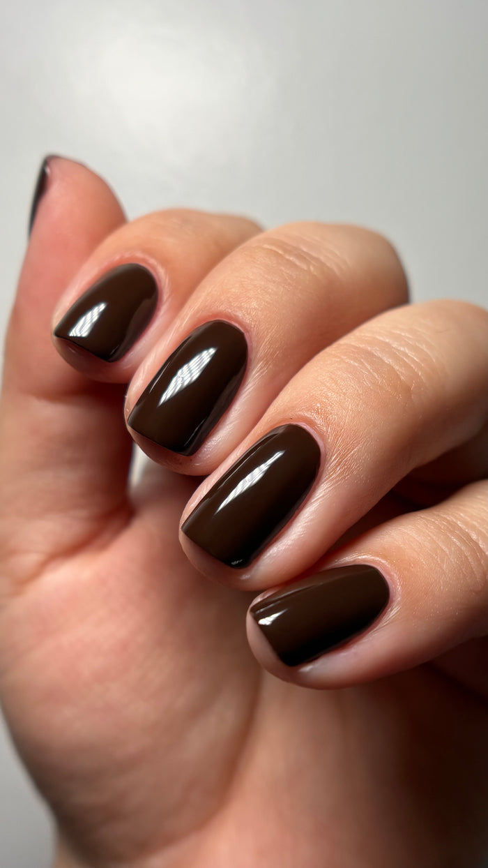 A hand with short, neatly manicured nails painted in LONDONTOWN’s glossy rich chocolate brown “Double Fudge” polish against a plain light background.