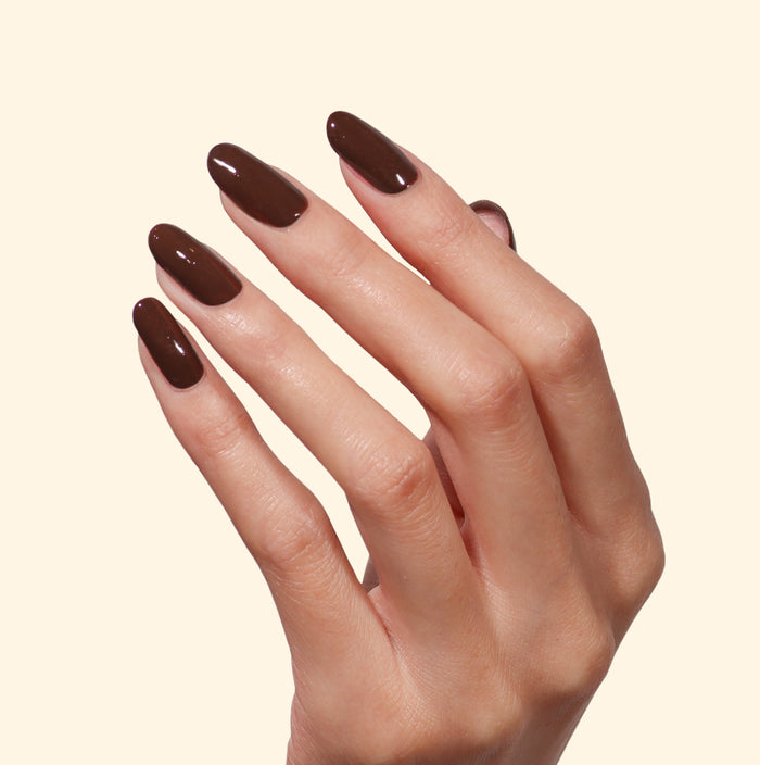 A hand with long, almond-shaped nails painted in LONDONTOWN’s Double Fudge, a rich chocolate brown, glossy nail polish, against a plain light background.