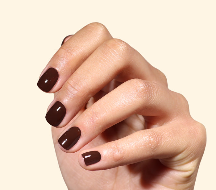 A hand with medium skin tone displays short, neatly manicured nails painted in LONDONTOWN’s Double Fudge—a rich, glossy chocolate brown polish—against a plain light background.