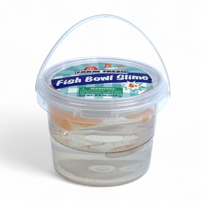 Farm Fresh Fish Bowl Slime by Farm Fresh comes in a clear plastic tub with handle, contains transparent slime with small colorful objects resembling goldfish, and features a warning plus net weight of 9.5 oz (269 g) on the label.