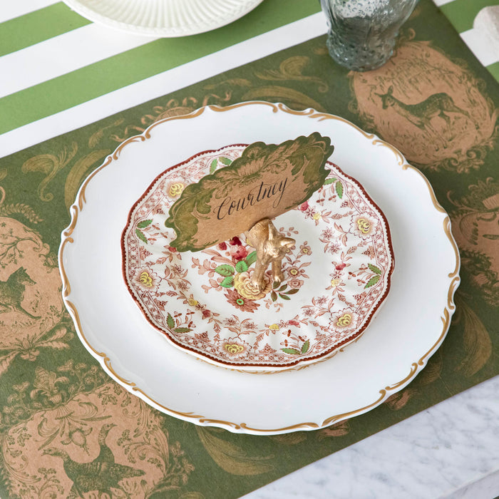 The Fox Name Card holder holding the Moss Fable Toile Place Card with "Courtney" written in cursive, held by a slot in the Fox's back, on a table setting with the Moss Fable Toile Placemat and Moss Green Classic Stripe Runner underneath.