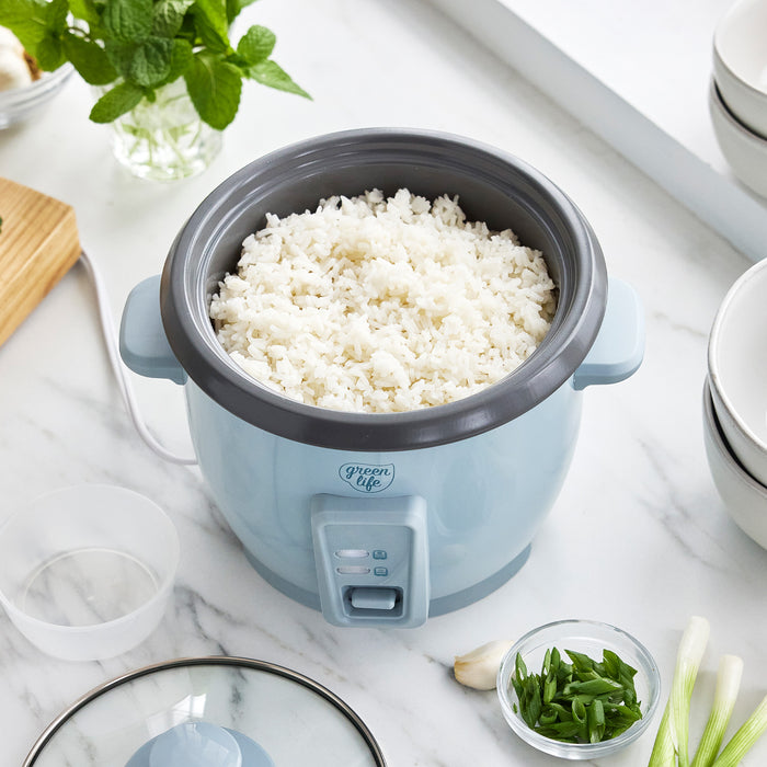 Blue 6-cup rice cooker filled with rice on a white countertop, next to bowls and fresh herbs.