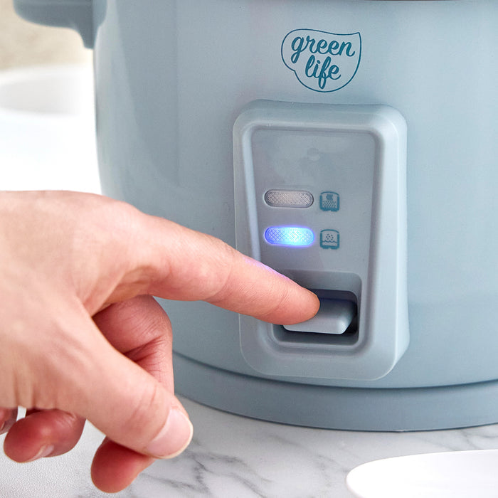Hand pressing the switch on a blue GreenLife 6-cup rice cooker with indicator lights above the button.
