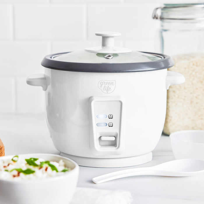 White 6-cup rice cooker with glass lid on counter, shown with a bowl of rice and a white spoon beside it.