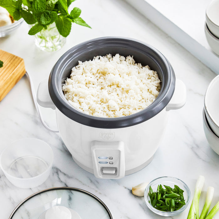 White 6-cup rice cooker on a marble counter, filled with rice, with bowls and fresh herbs nearby.