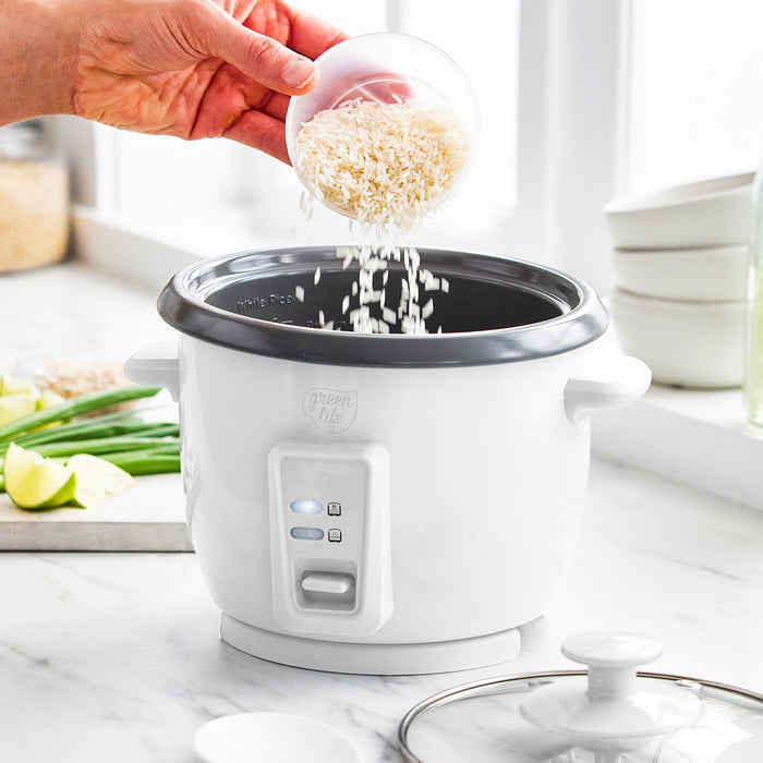 Hand pouring uncooked rice into a white GreenLife 6-cup rice cooker on kitchen counter with assorted vegetables nearby.