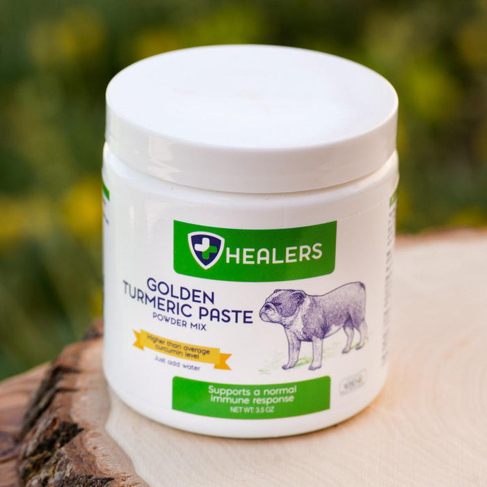 White jar of Healers Golden Turmeric Paste powder mix. Label shows bulldog and text about immune support. Jar is on light wood, with blurred natural background.