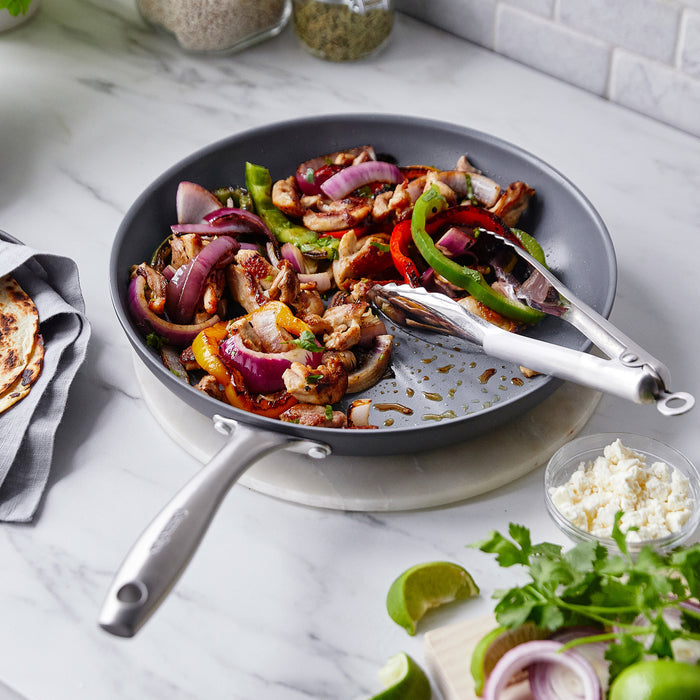 Chicken, peppers, and onions cooking in a 10 ceramic frypan with bamboo spatula, topped with lime wedges and cheese.