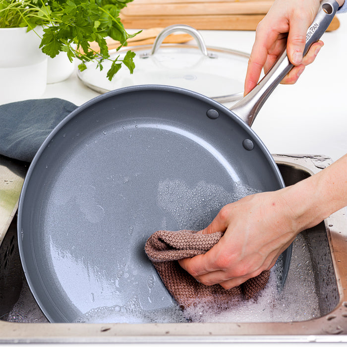 Washing a ceramic nonstick frypan with a brown sponge in a soapy sink; bamboo spatula and plants nearby.