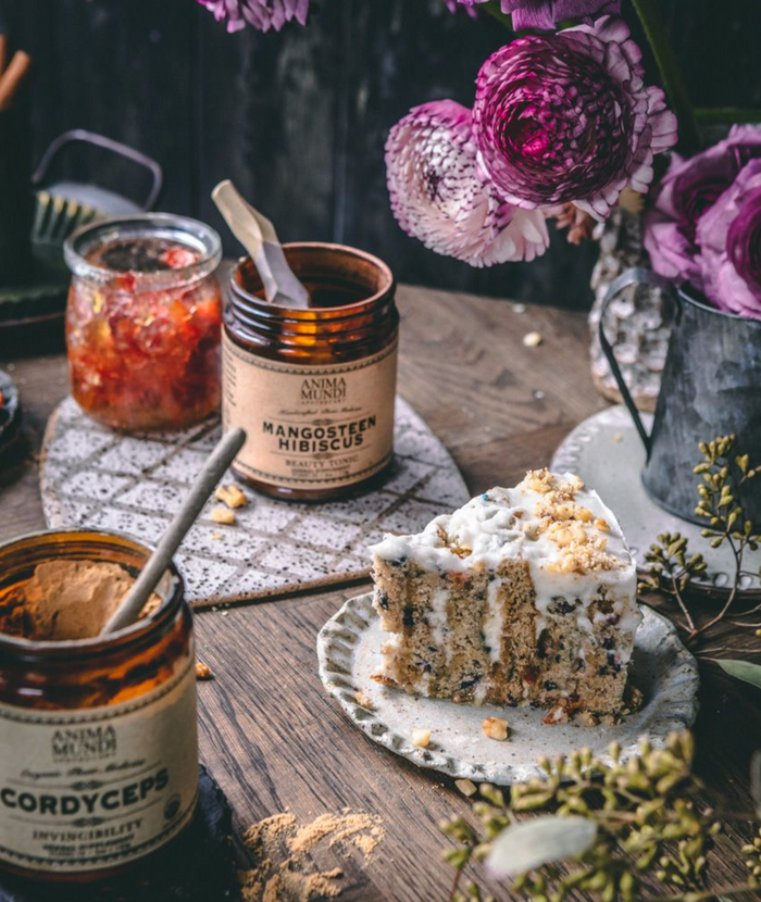 Jars of 'Anima Mundi' cordyceps and manna honey on a wooden table with a slice of cake and flowers.