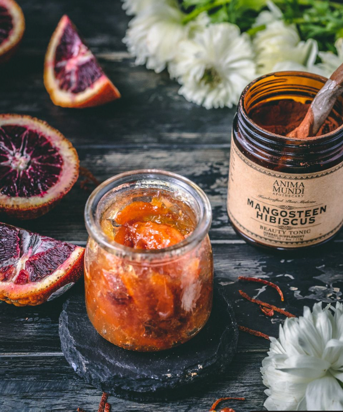 Two jars of a fruit preserve on a dark surface with blood oranges and white flowers.