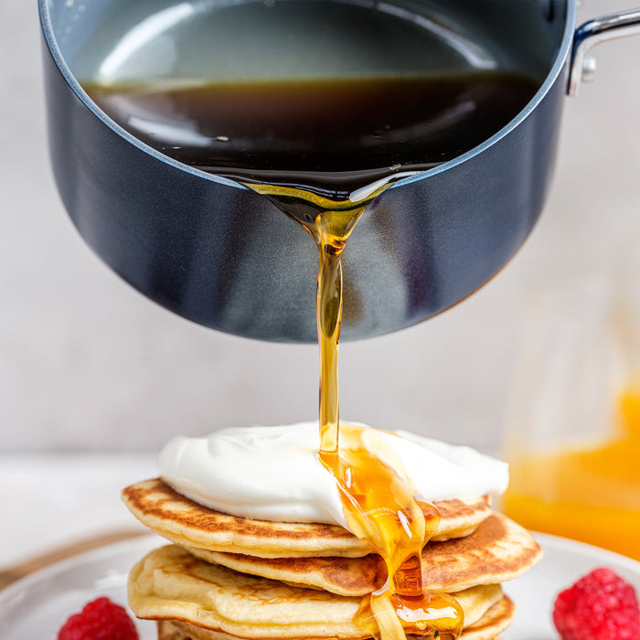 Navy ceramic nonstick saucepan pouring syrup onto pancakes topped with cream and raspberries.
