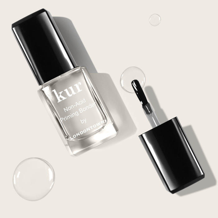 A bottle of LONDONTOWN Non-Acid Priming Bonder with a black cap is shown. The cap is removed, revealing a brush applicator. Clear droplets are scattered nearby—a pro secret for making your manis last longer.