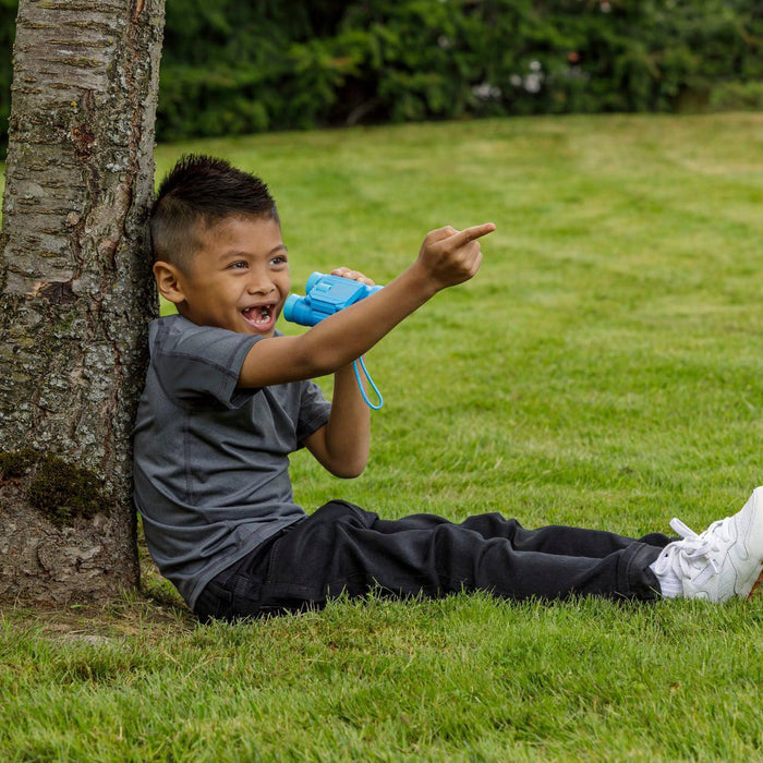 A young boy sits on the grass, leaning against a tree and smiling as he points and holds an Outdoor Discovery Folding Binocular by Outdoor Discovery, surrounded by trees—perfect for outdoor exploration.