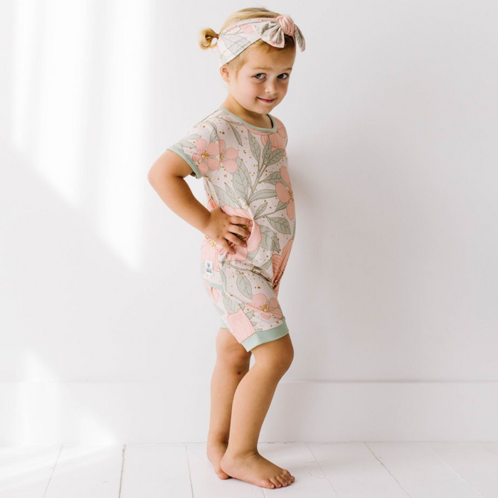 A toddler wearing a floral shorty romper in pink and green colors, with a matching headband.