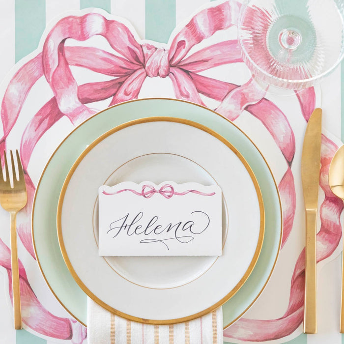 A place setting with a plate and gold cutlery, adorned with a Hester & Cook Pink Bow Place Card for added loveliness.