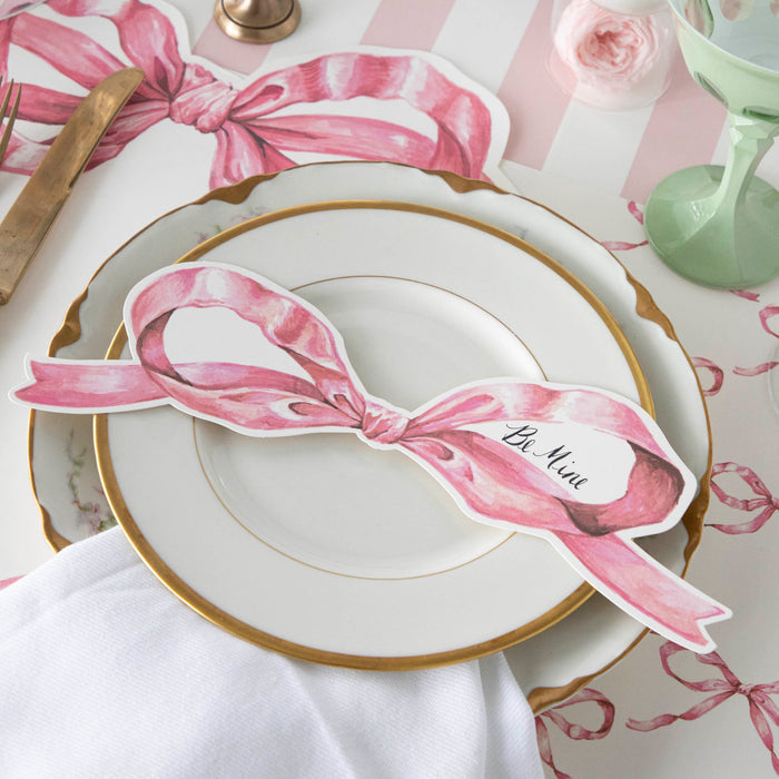 A Pink Bow Table Accent with "Be Mine" written in the blank space in the right bow loop resting on the plate of a pink place setting.