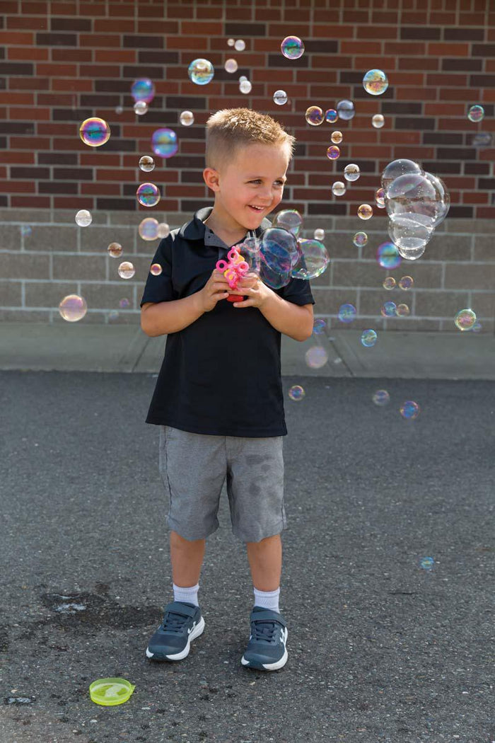 A young boy enjoys outdoor fun, smiling as he uses the Playground Classics Turbo Bubble Blower to create a flurry of colorful bubbles. Dressed in a black polo, gray shorts, and sneakers, he stands on pavement with a brick wall behind him.