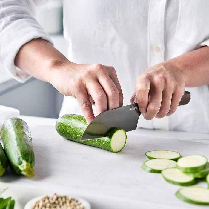 Zucchini being sliced on a white cutting board with a small Santoku knife featuring a sharp blade and walnut handle.