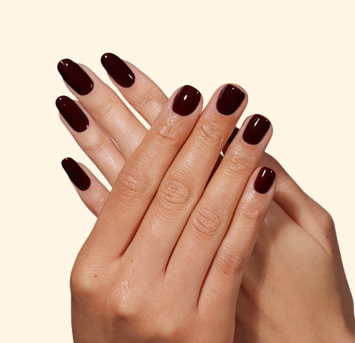 Two hands with neatly manicured nails painted in LONDONTOWN’s Red Velvet, a glossy deep burgundy polish, fingers gently crossed against a plain light beige background.
