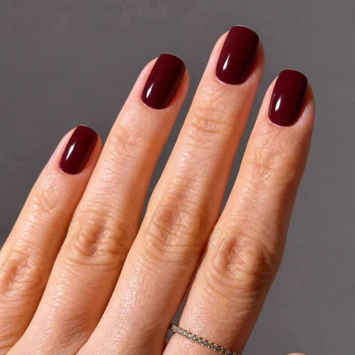A hand with short, neatly manicured nails painted in LONDONTOWN’s “Red Velvet” shade is shown against a plain gray background. The ring finger is adorned with a thin, silver ring.