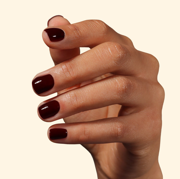 A hand with neatly manicured short nails painted in LONDONTOWN’s Red Velvet, a glossy deep burgundy shade, held against a plain light background.
