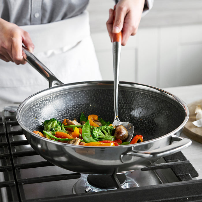 Person stirring vegetables in a ceramic nonstick wok with glass lid on a gas stove.