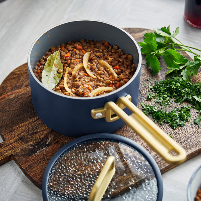 Blue 2-quart saucepan with lentils, onions, and bay leaf next to chopped parsley on a wooden board.