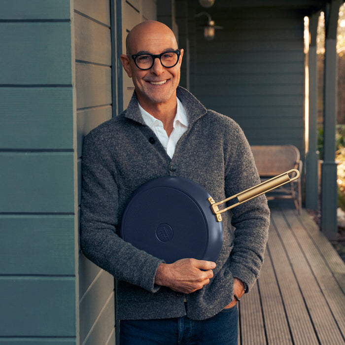 Man in glasses and gray sweater holding a blue ceramic nonstick frypan outdoors.