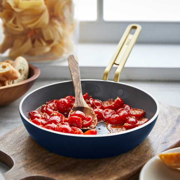 Blue ceramic nonstick frypan with cherry tomatoes being stirred by a wooden spoon, pasta nearby.