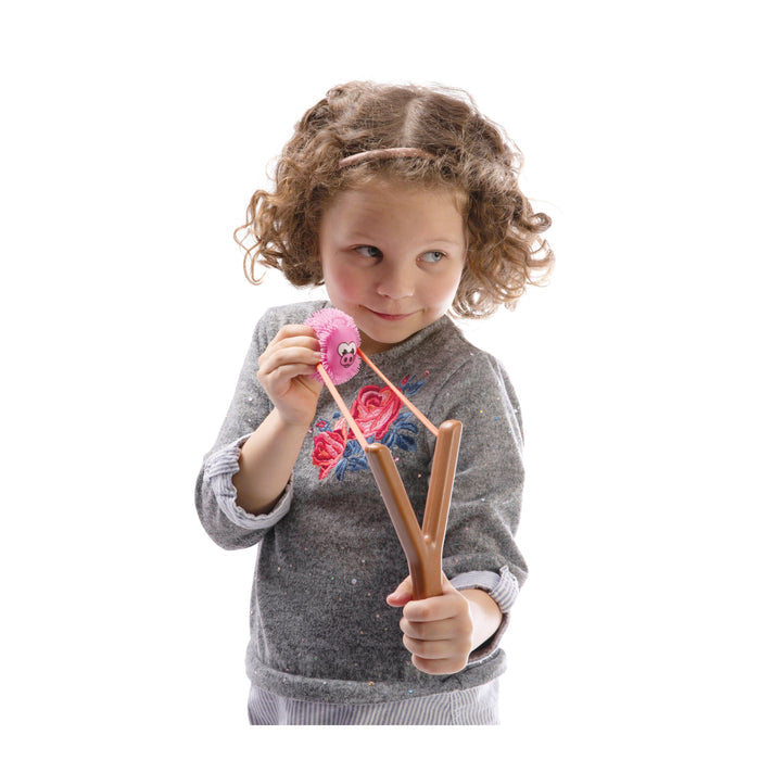 A young child in a gray sweater playfully aims the YAY! When Pigs Fly novelty slingshot by YAY!, featuring a humorous pink flying pig, while looking to the side against a white background.